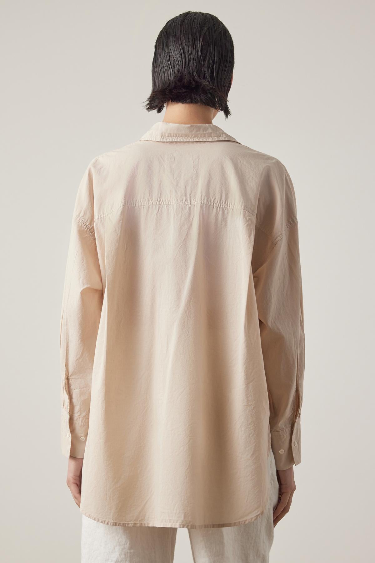 A person with dark hair, seen from behind, wearing an oversized REDONDO BUTTON-UP SHIRT made of soft cotton by Velvet by Jenny Graham and light-colored relaxed trousers, standing against a plain background.-37618953584833