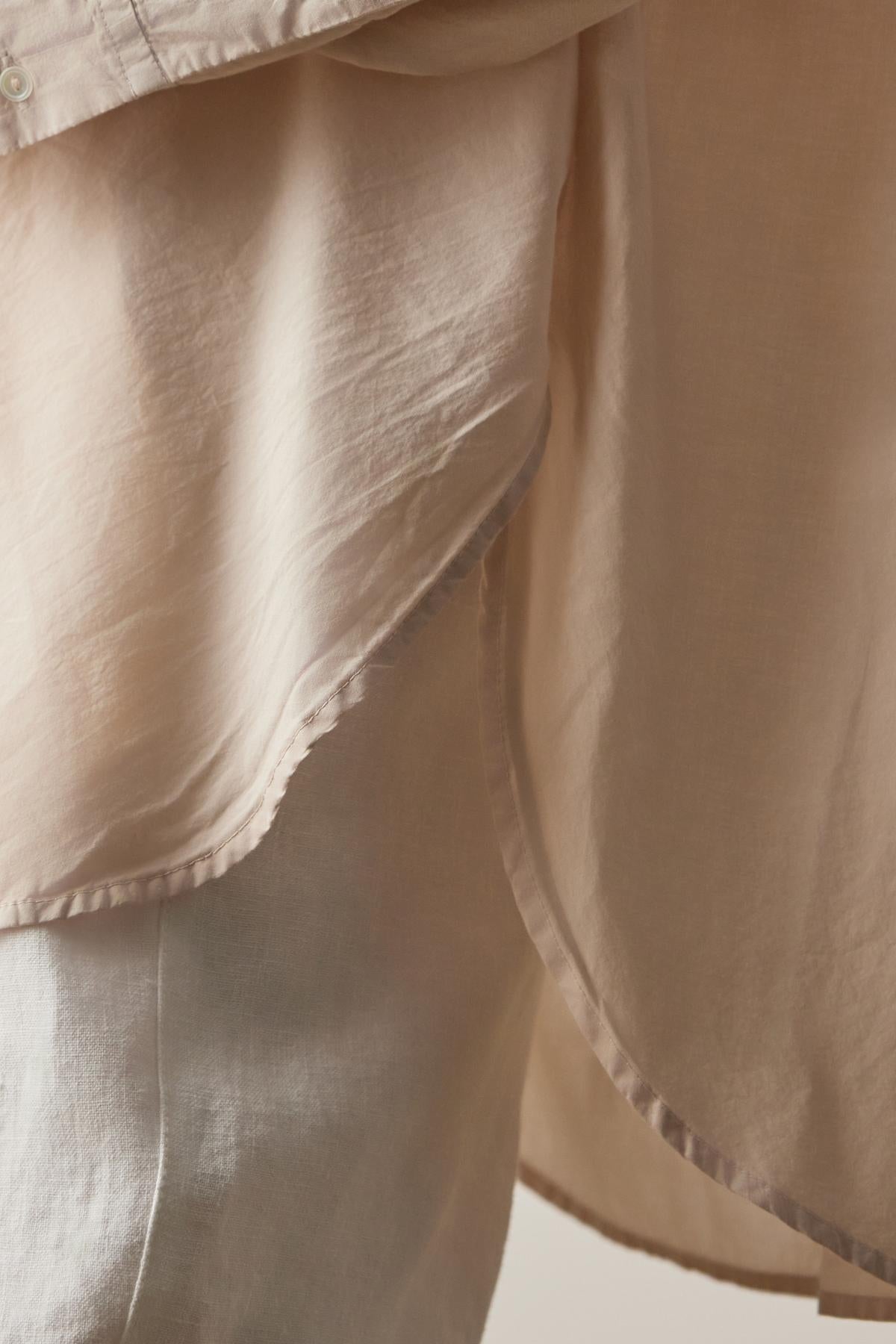   Close-up of a light beige, semi-transparent fabric overlapping and draping, showcasing its smooth texture and delicate layers, reminiscent of the soft cotton used in the REDONDO BUTTON-UP SHIRT by Velvet by Jenny Graham. 