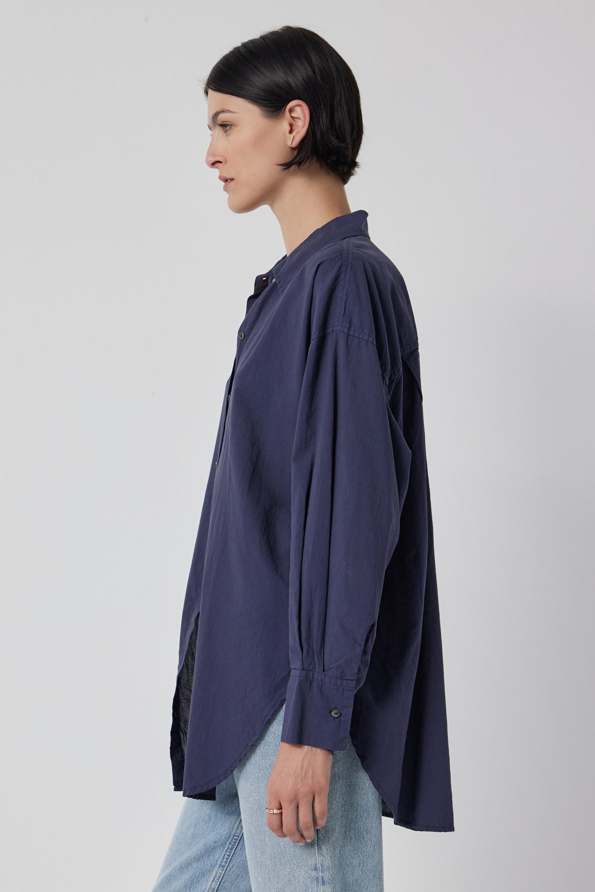 A side profile of a person wearing an oversized navy blue REDONDO button-up shirt made from soft cotton shirting and light blue relaxed trousers against a neutral background by Velvet by Jenny Graham.-36168814657729