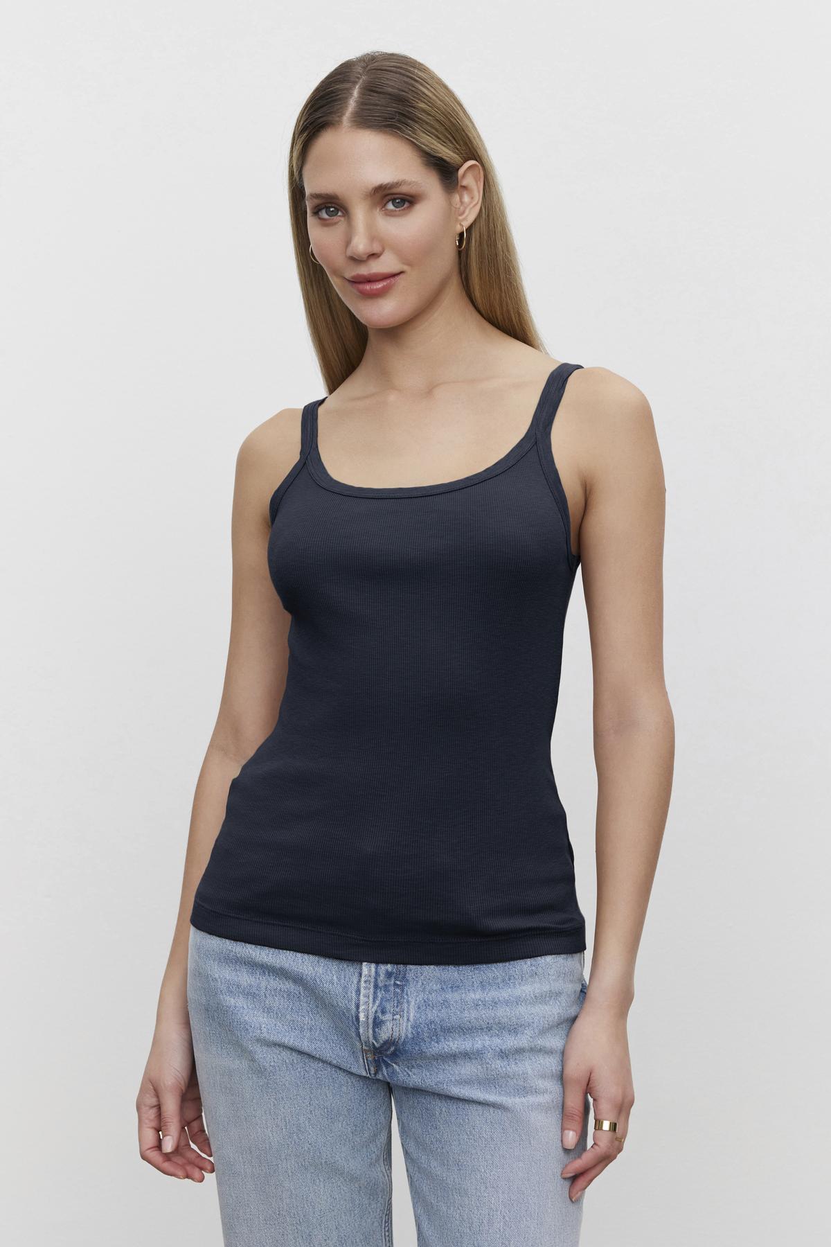   A woman with brown hair, standing in a white background, wearing a navy Aliza Ribbed Cotton Layering Tank by Velvet by Graham & Spencer with double-layer straps and light blue jeans. 