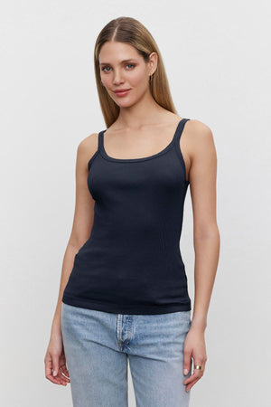 A woman with brown hair, standing in a white background, wearing a navy Aliza Ribbed Cotton Layering Tank by Velvet by Graham & Spencer with double-layer straps and light blue jeans.