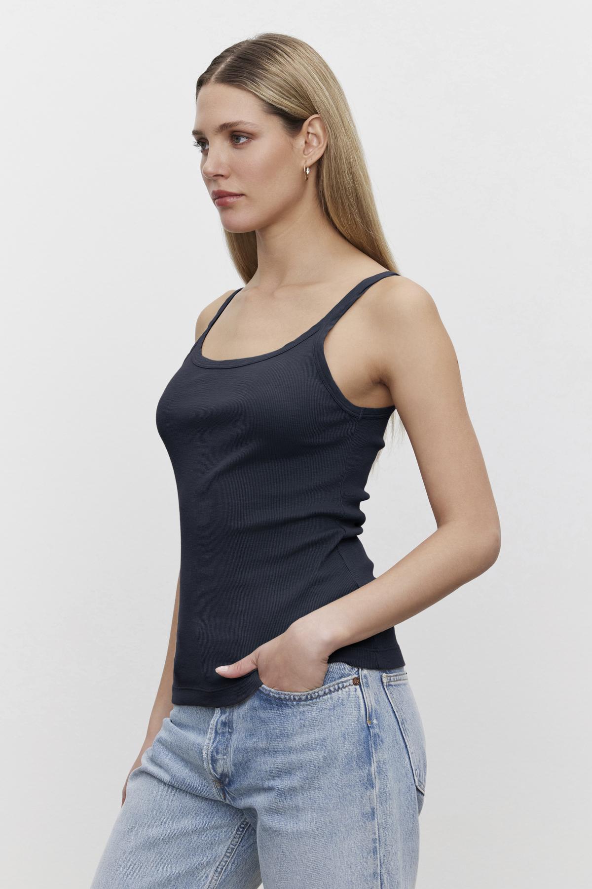 A woman in a Velvet by Graham & Spencer ALIZA RIBBED COTTON LAYERING TANK with double-layer straps and light blue jeans standing against a white background, looking to the side.-36580672864449