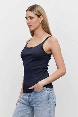 A woman in a Velvet by Graham & Spencer ALIZA RIBBED COTTON LAYERING TANK with double-layer straps and light blue jeans standing against a white background, looking to the side.