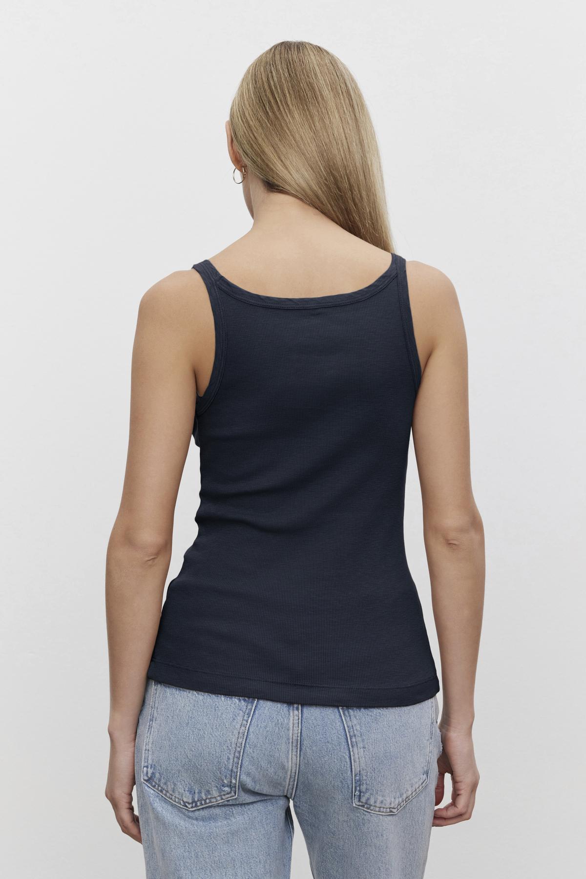   A woman viewed from behind wearing a Velvet by Graham & Spencer ALIZA RIBBED COTTON LAYERING TANK with double-layer straps and light blue jeans, standing against a plain white background. 