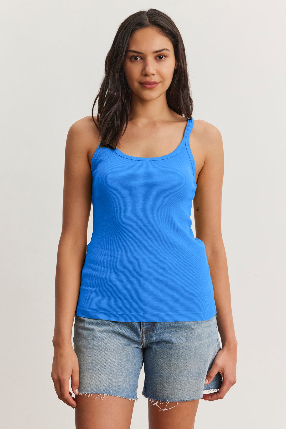 A woman stands facing forward in the Velvet by Graham & Spencer ALIZA RIBBED COTTON LAYERING TANK and light denim shorts against a plain light background.-39149199163585