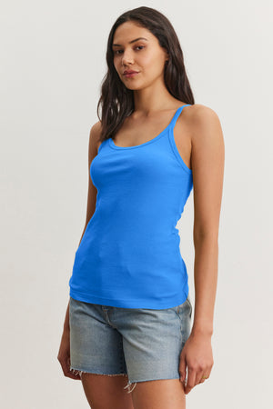 A woman wears the ALIZA RIBBED COTTON LAYERING TANK by Velvet by Graham & Spencer in bright blue with denim shorts, standing against a plain light background—ideal for easy layering.
