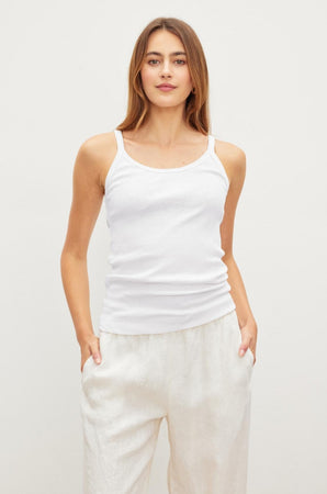 A woman with long hair is wearing an ALIZA RIBBED COTTON LAYERING TANK by Velvet by Graham & Spencer with double-layer straps and light-colored pants, standing with her hands in her pockets against a plain background.