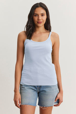 A woman with long dark hair is wearing a light blue ALIZA RIBBED COTTON LAYERING TANK from Velvet by Graham & Spencer and denim shorts. She is standing against a plain white background.