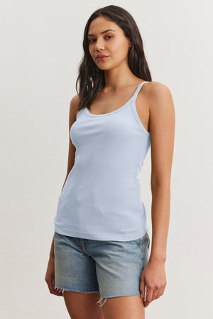 A person with long hair wearing a light blue ALIZA RIBBED COTTON LAYERING TANK by Velvet by Graham & Spencer and denim shorts stands against a plain background.