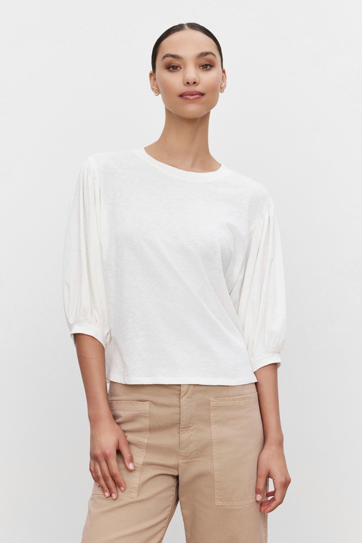   A person stands against a plain white background, wearing the AMARA TEE by Velvet by Graham & Spencer with banded cuffs and beige pants. Their hair is pulled back. 