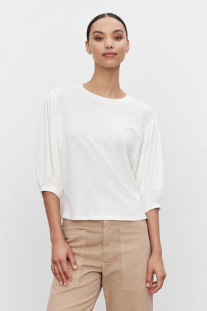 A person stands against a plain white background, wearing the AMARA TEE by Velvet by Graham & Spencer with banded cuffs and beige pants. Their hair is pulled back.