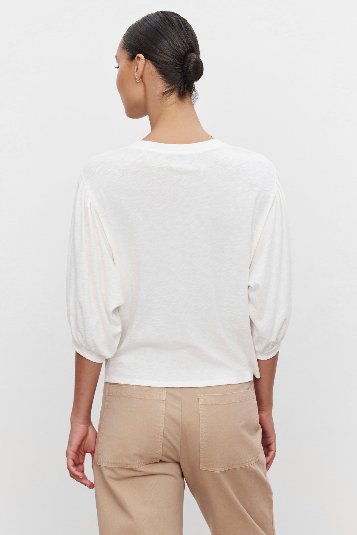   A person is shown from the back, wearing a white long-sleeved AMARA TEE with banded cuffs by Velvet by Graham & Spencer and light brown pants. The person's hair is tied back in a neat bun. 