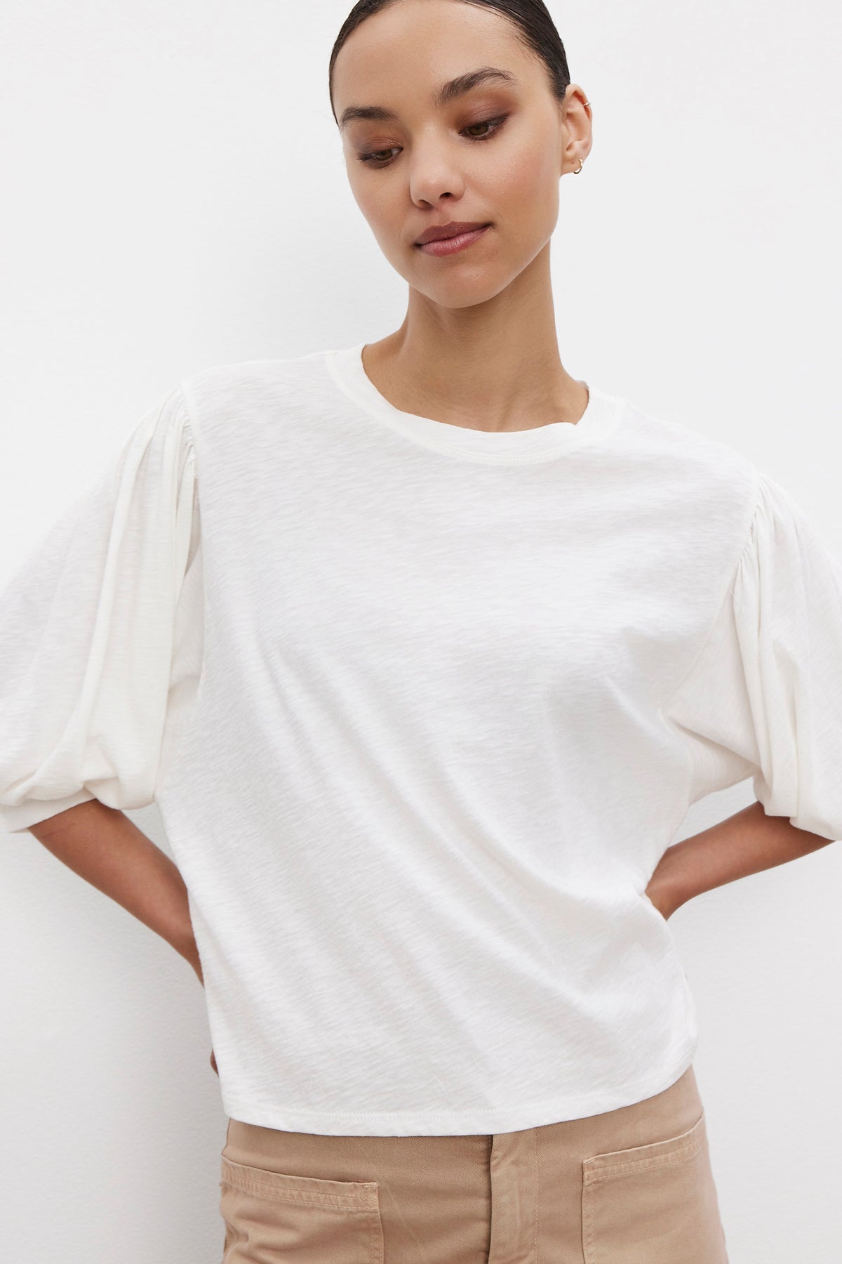   A person stands against a plain background, wearing the AMARA TEE by Velvet by Graham & Spencer—a white short-sleeve top with extra volume sleeves—and tan pants. Their hands are placed on their hips. 