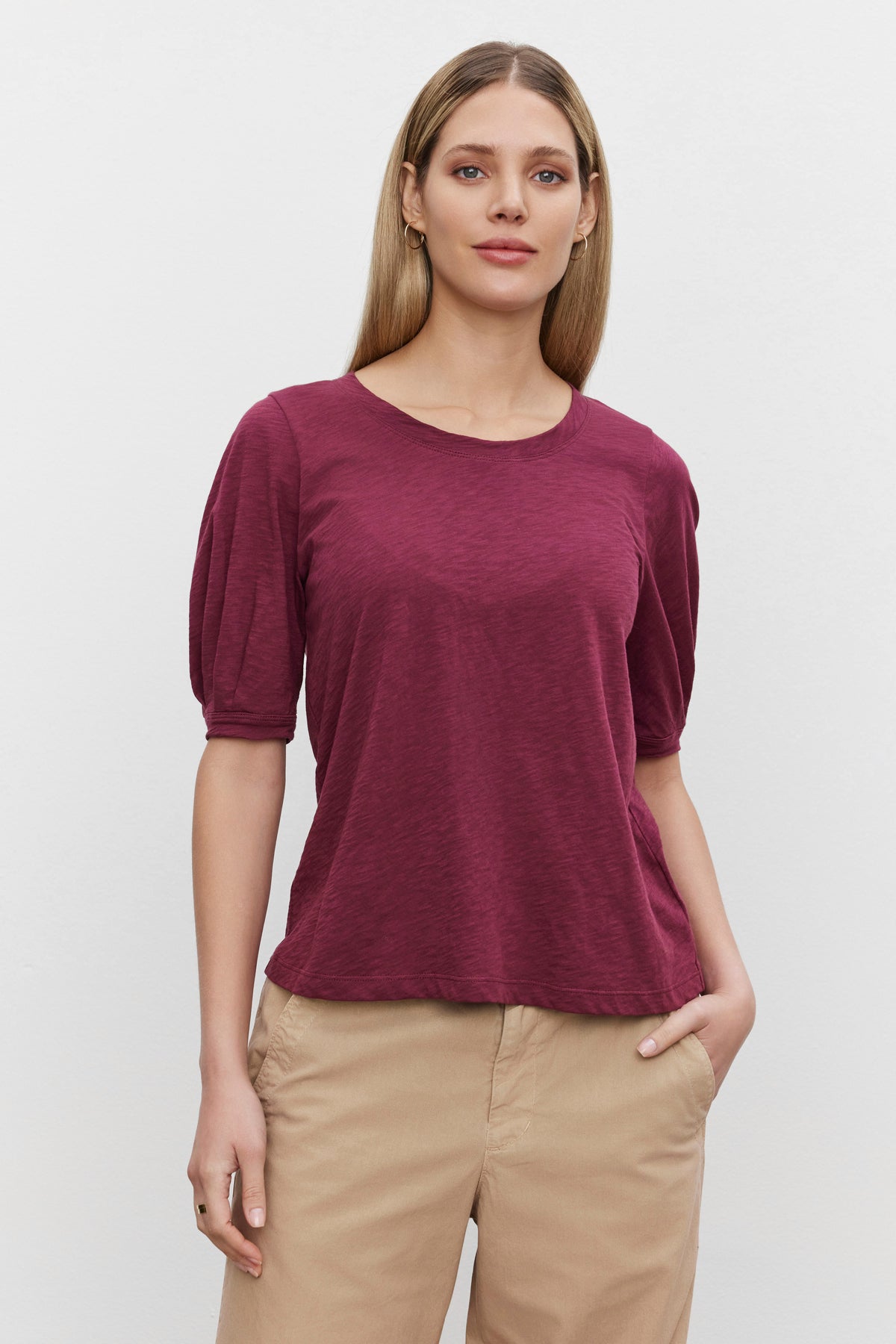   A person with long hair is standing against a plain white background, wearing a maroon ANABELLE TEE by Velvet by Graham & Spencer and beige pants. 