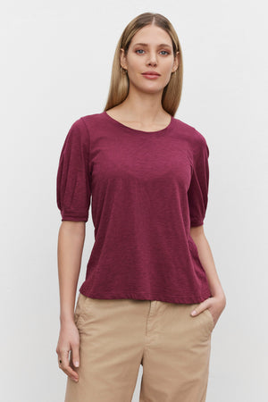 A person with long hair is standing against a plain white background, wearing a maroon ANABELLE TEE by Velvet by Graham & Spencer and beige pants.