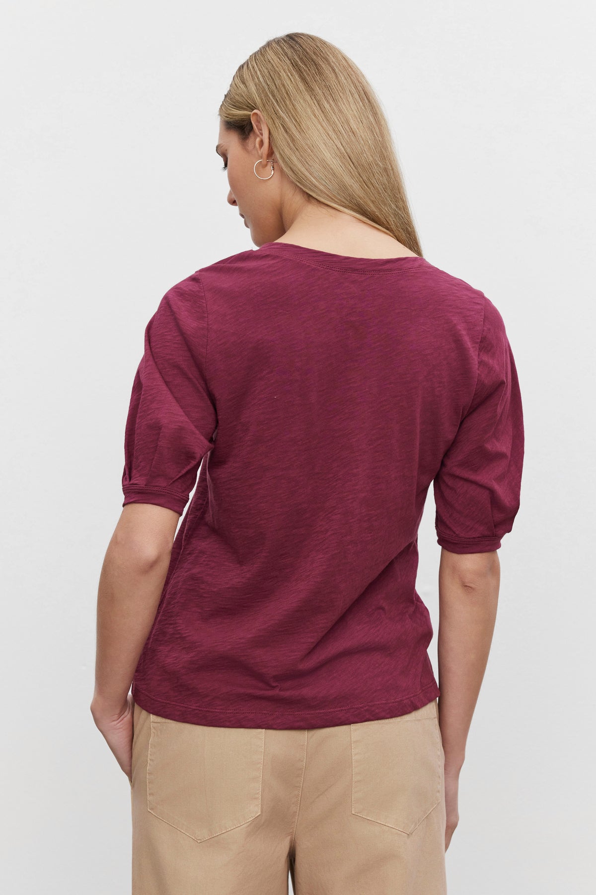   A person with long blonde hair stands facing away, wearing a maroon Velvet by Graham & Spencer ANABELLE TEE and beige pants against a plain white background. 