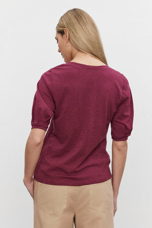 A person with long blonde hair stands facing away, wearing a maroon Velvet by Graham & Spencer ANABELLE TEE and beige pants against a plain white background.