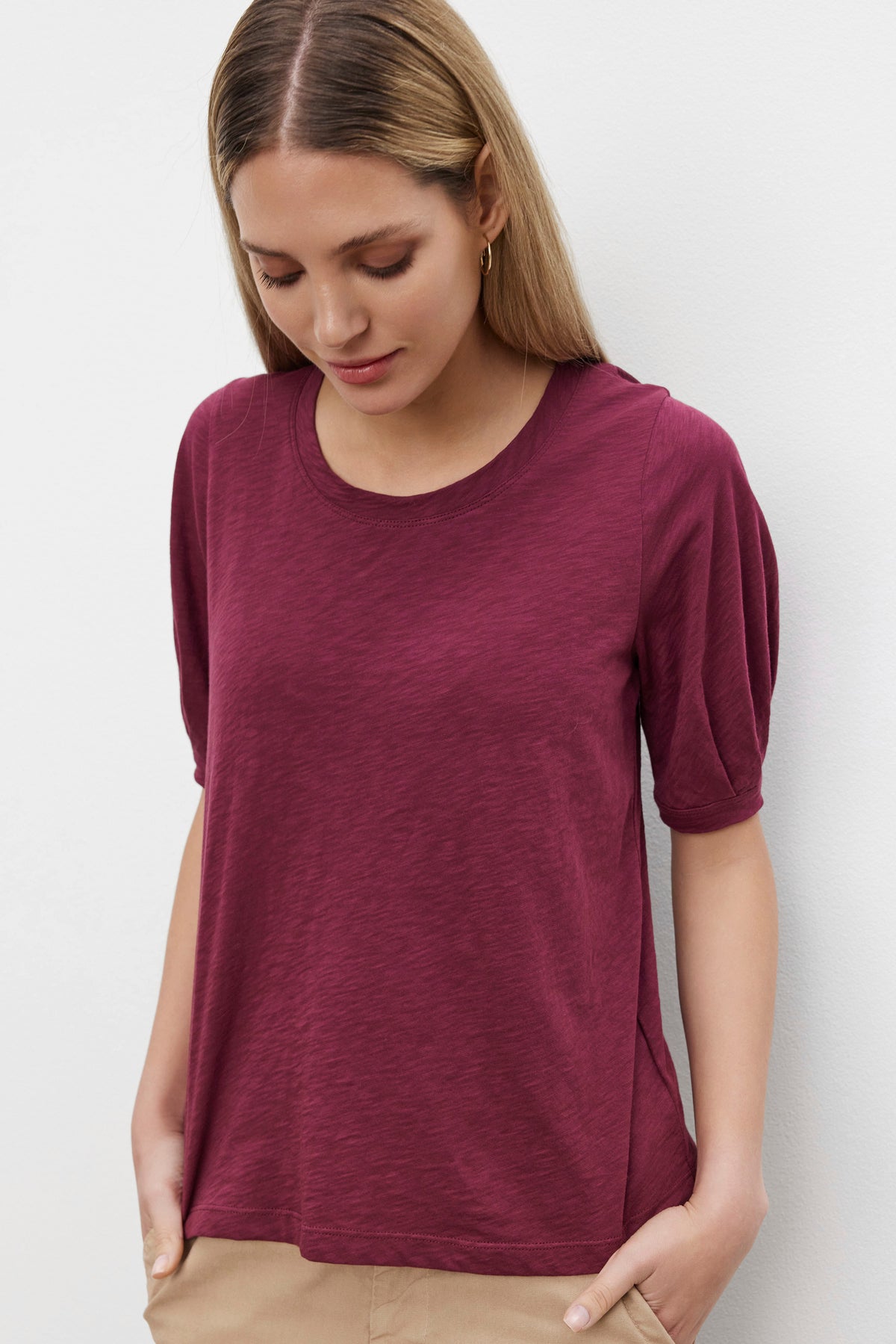   A woman with long, blonde hair, wearing the ANABELLE TEE by Velvet by Graham & Spencer in maroon, looks down with her hands in the pockets of her beige pants. 