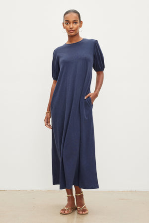 A black woman in a Velvet by Graham & Spencer ASTRID cotton slub maxi dress with puff sleeves, standing against a white background. She wears hoop earrings and brown sandals.