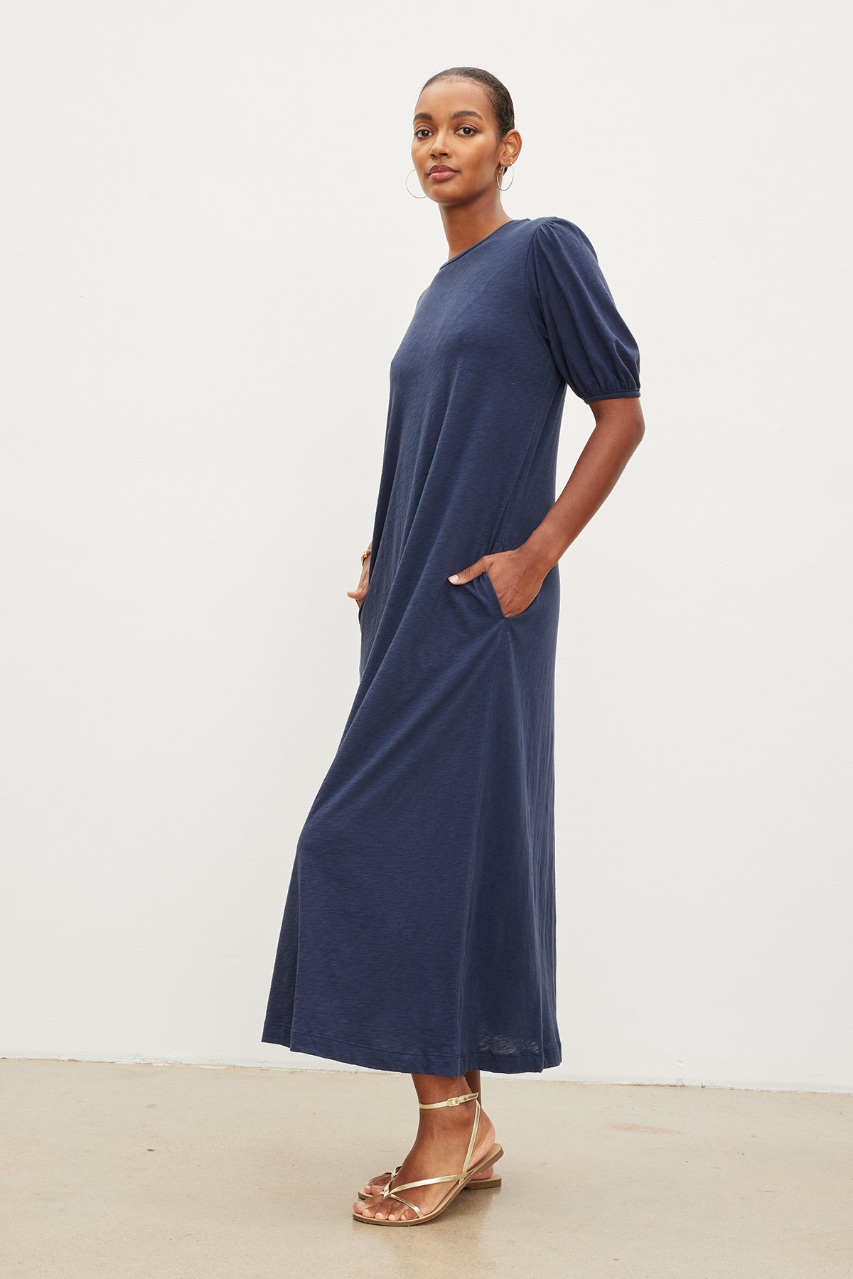   A woman in a navy blue Astrid Cotton Slub Maxi Dress with puff sleeves and gold sandals stands confidently with one hand on her hip in a white studio. Brand name: Velvet by Graham & Spencer. 