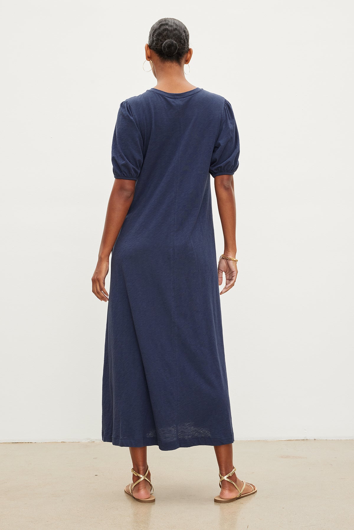   A woman in an Astrid Cotton Slub Maxi Dress by Velvet by Graham & Spencer with puff sleeves, viewed from the back, standing against a plain white backdrop. She's wearing gold sandals. 