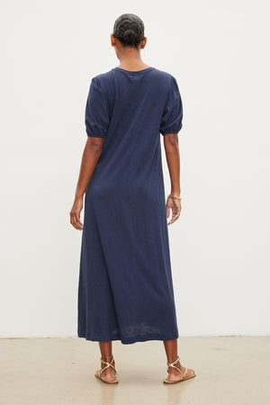 A woman in an Astrid Cotton Slub Maxi Dress by Velvet by Graham & Spencer with puff sleeves, viewed from the back, standing against a plain white backdrop. She's wearing gold sandals.