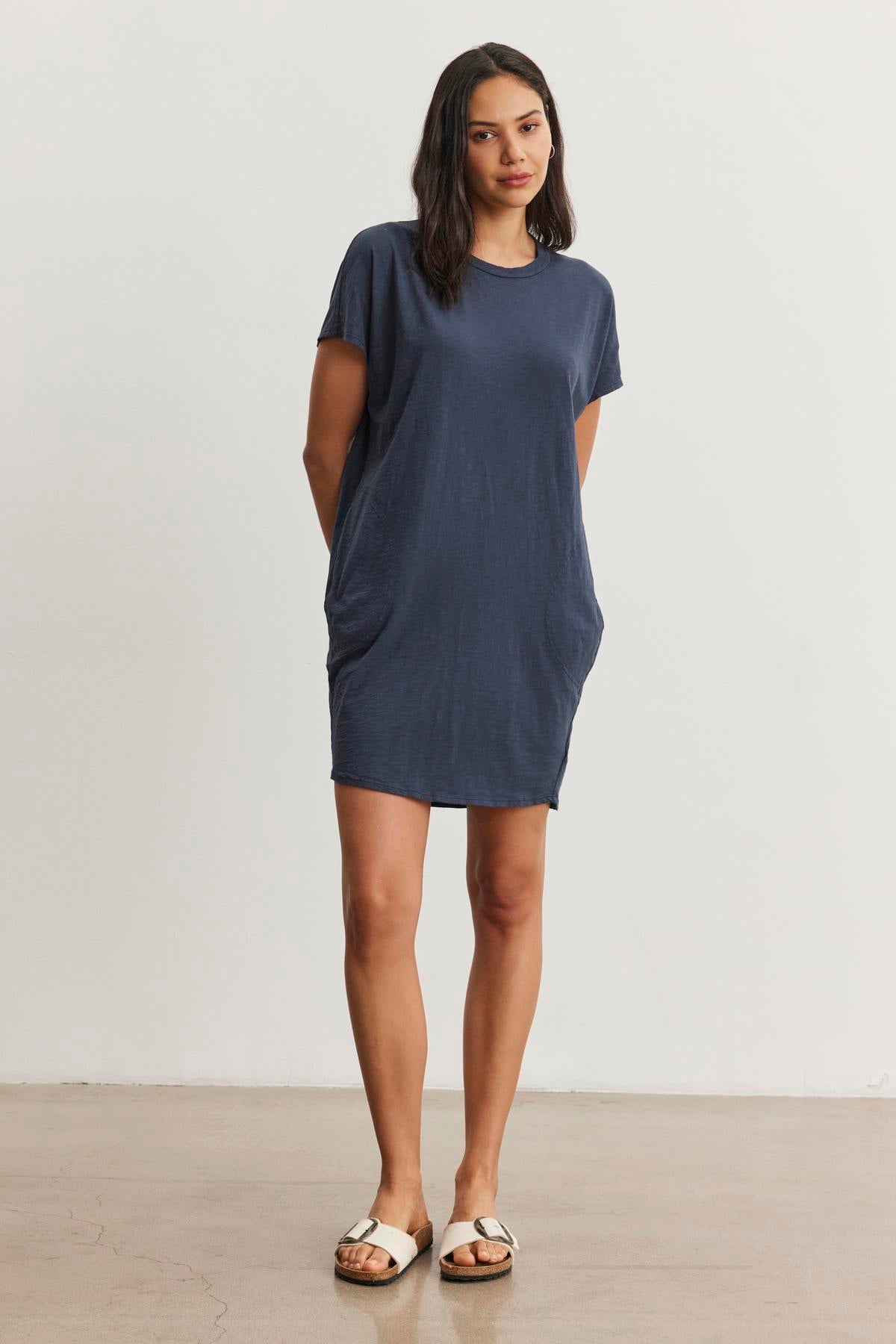   A person with long hair stands wearing a relaxed fit, short-sleeve blue ATHENA DRESS by Velvet by Graham & Spencer with hands in pockets and white sandals, against a plain white background. 