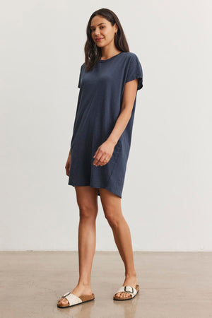 A woman stands against a plain background wearing a short-sleeved navy blue ATHENA DRESS by Velvet by Graham & Spencer and white sandals. She has long, dark hair and a neutral expression, effortlessly showcasing the relaxed fit of her outfit.