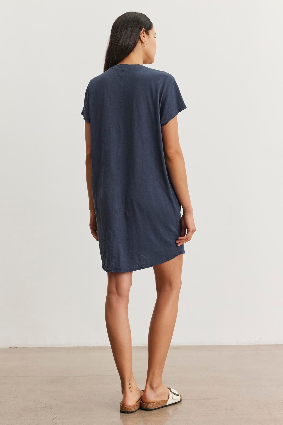 A woman stands facing away from the camera, wearing the ATHENA DRESS by Velvet by Graham & Spencer, a relaxed fit dark blue t-shirt dress made of cotton slub fabric, and white sandals.-37606520291521