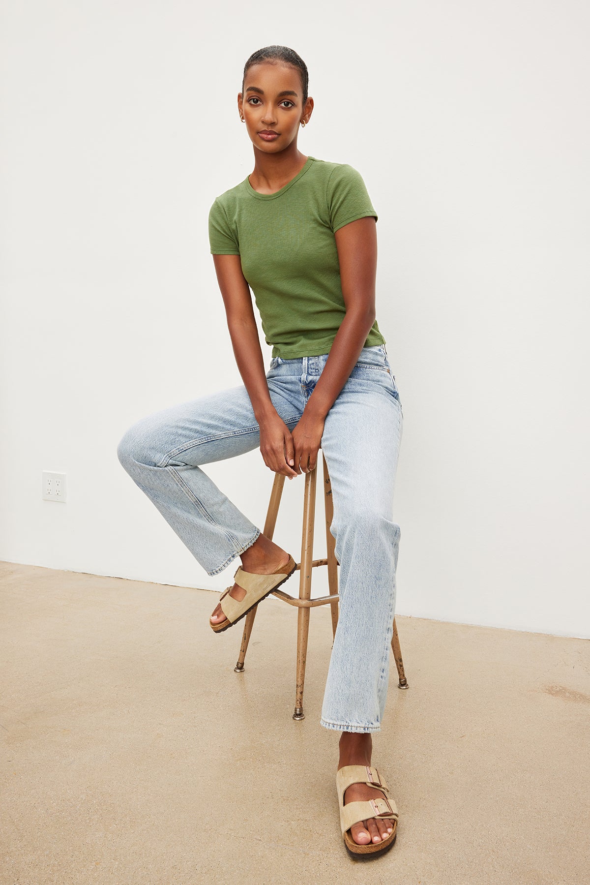 A woman is sitting on a stool in a cozy Velvet by Graham & Spencer BRENNY RIBBED CREW NECK TEE made of ribbed cotton knit.-35955657932993