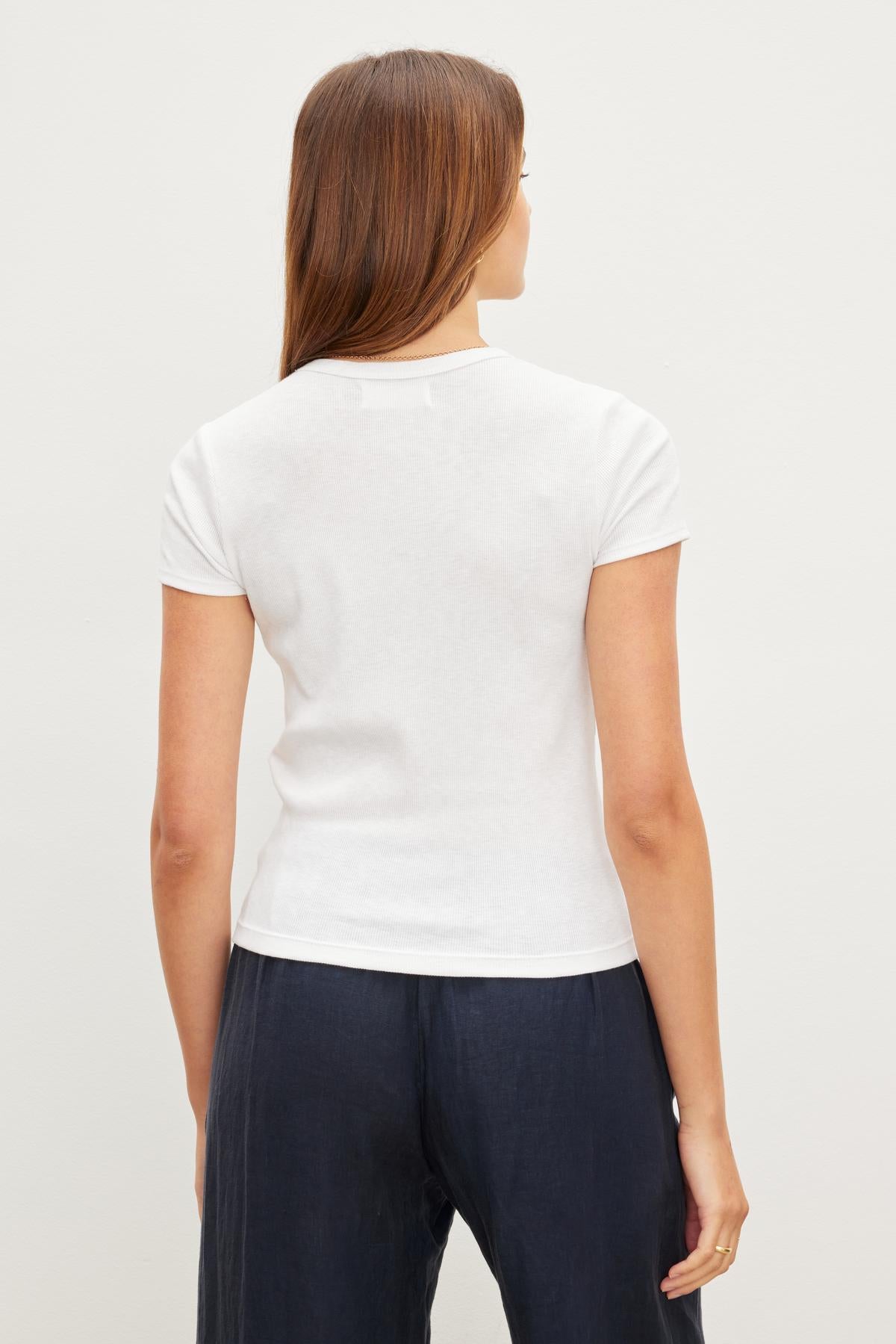   The back view of a woman wearing a slightly cropped white BRENNY RIBBED CREW NECK TEE, crafted from ribbed cotton knit for a cozy feel, by Velvet by Graham & Spencer. 