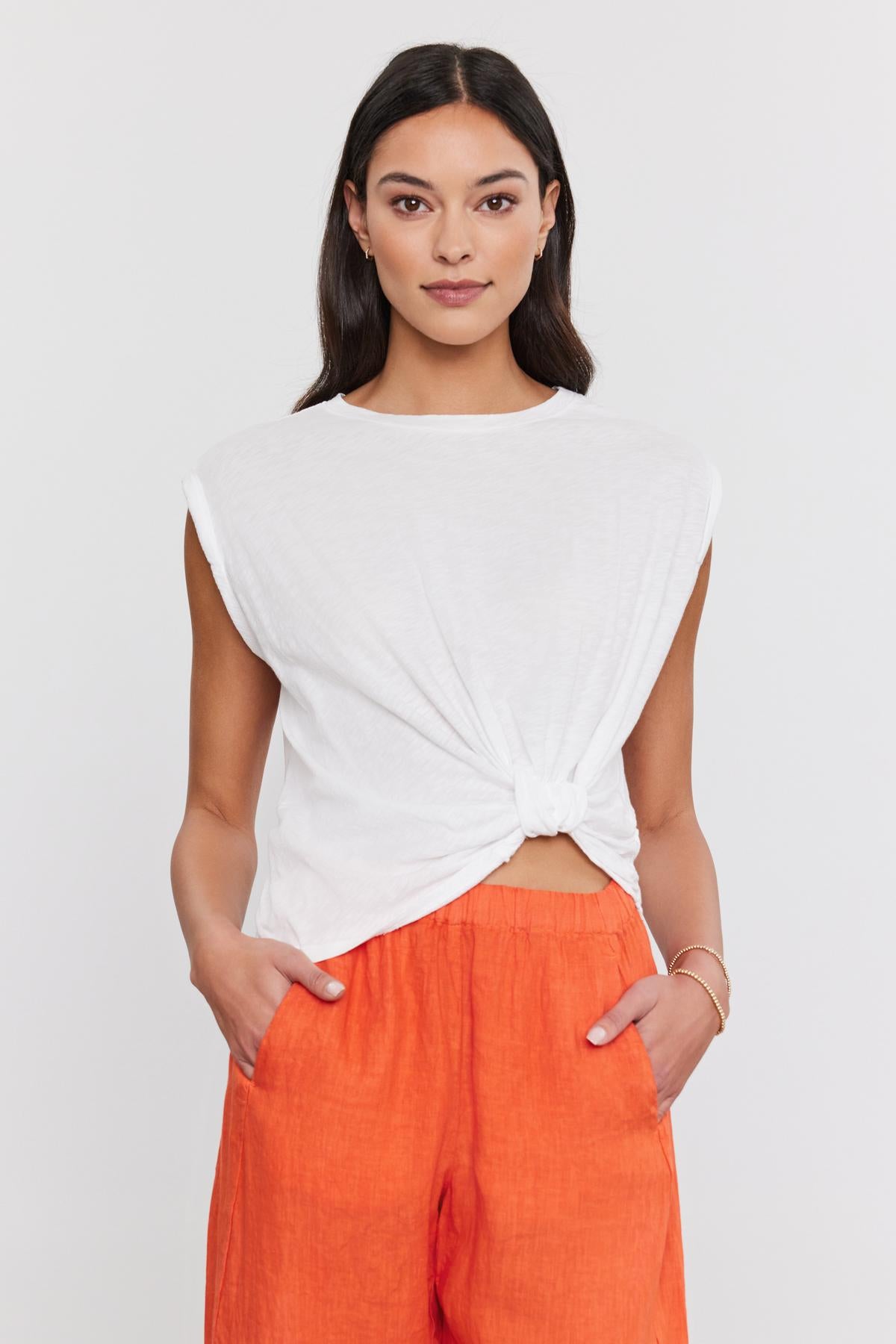   Woman standing, wearing a white CALLISTA TEE by Velvet by Graham & Spencer with a twist knot detail and orange pants, looking directly at the camera with a slight smile. 