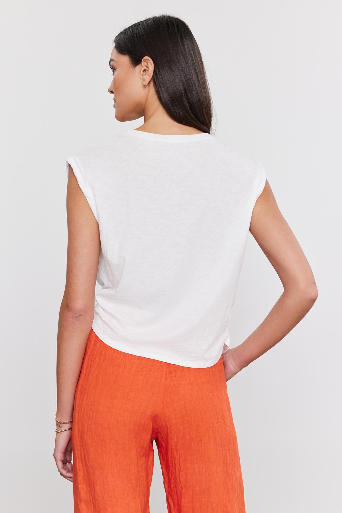   A woman viewed from behind, wearing a white cotton slub CALLISTA TEE by Velvet by Graham & Spencer with a crew neckline and orange pants, standing against a plain background. 