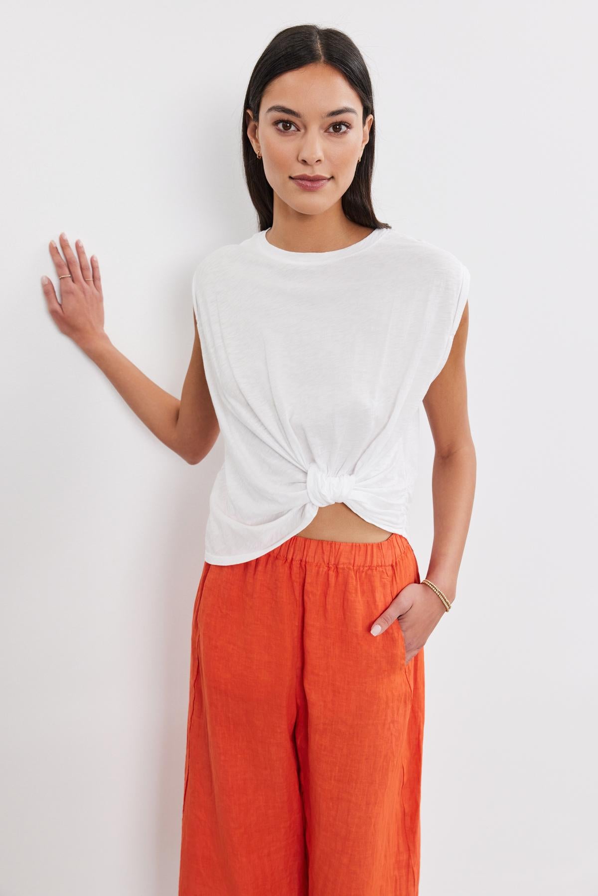   A woman in a Velvet by Graham & Spencer CALLISTA TEE with twist knot detail and orange pants standing against a white background, with one hand on her hip and the other raised slightly. 