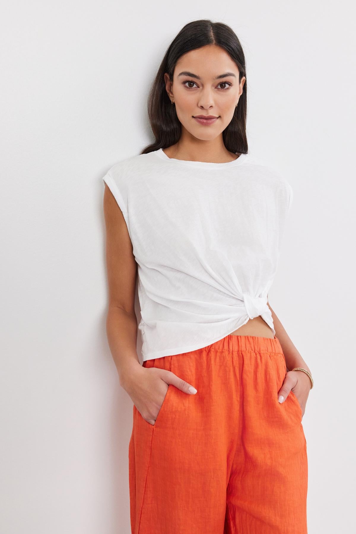   A woman in a white Velvet by Graham & Spencer CALLISTA TEE with a crew neckline and bright orange trousers, standing against a white background, looking at the camera. 