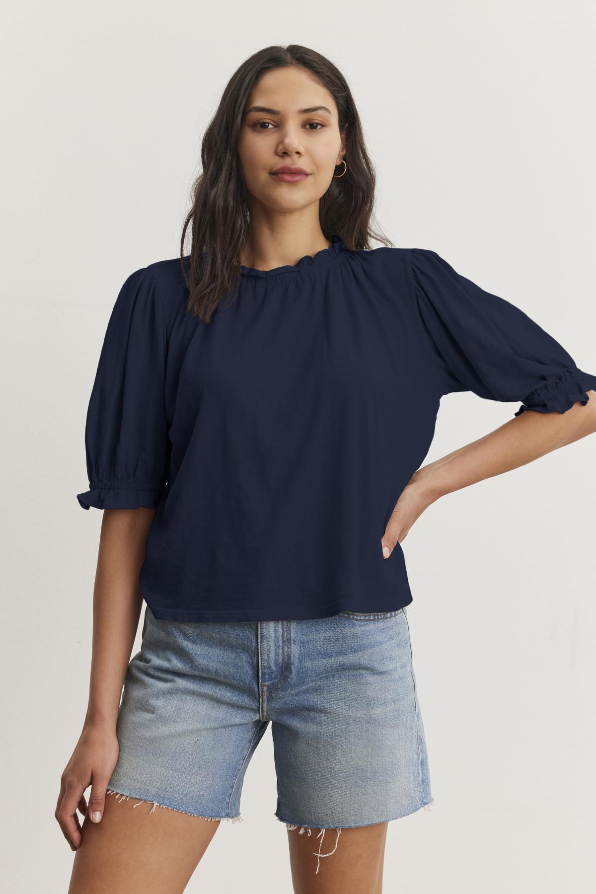   A person with long hair is wearing a navy blue Velvet by Graham & Spencer CARLENE TEE with puffed sleeves and an elastic neckline, made of soft cotton slub fabric, paired with denim shorts, standing against a plain white background. 