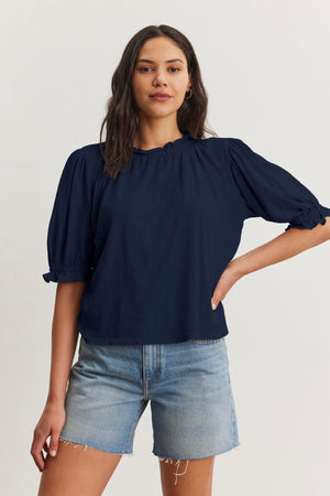 A person with long hair is wearing a navy blue Velvet by Graham & Spencer CARLENE TEE with puffed sleeves and an elastic neckline, made of soft cotton slub fabric, paired with denim shorts, standing against a plain white background.