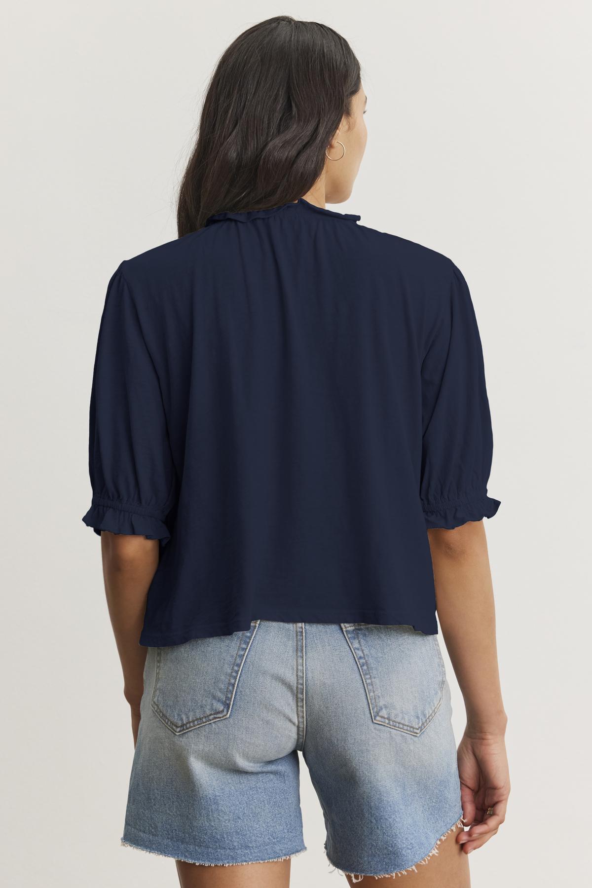   Back view of a person with long hair wearing a navy blue CARLENE TEE by Velvet by Graham & Spencer with short ruffled sleeves, an elastic neckline, and light blue denim shorts. 