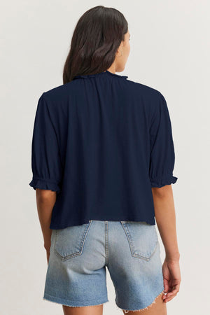 Back view of a person with long hair wearing a navy blue CARLENE TEE by Velvet by Graham & Spencer with short ruffled sleeves, an elastic neckline, and light blue denim shorts.