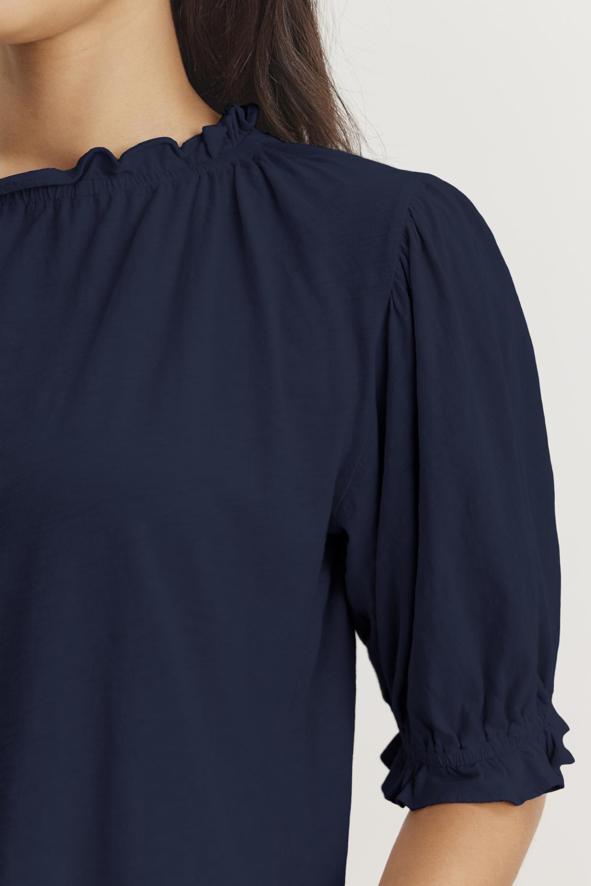   Close-up of a person wearing a dark blue CARLENE TEE from Velvet by Graham & Spencer made from cotton slub fabric, featuring gathered details along the ruffle elastic neckline and puffed, elbow-length sleeves. The background is plain and light-colored. 