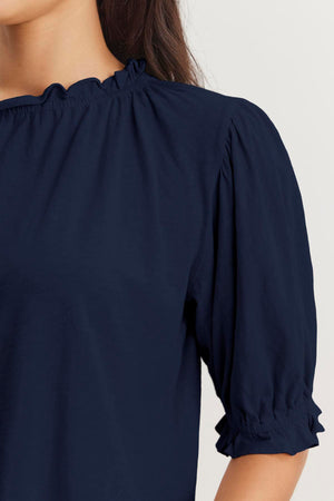 Close-up of a person wearing a dark blue CARLENE TEE from Velvet by Graham & Spencer made from cotton slub fabric, featuring gathered details along the ruffle elastic neckline and puffed, elbow-length sleeves. The background is plain and light-colored.