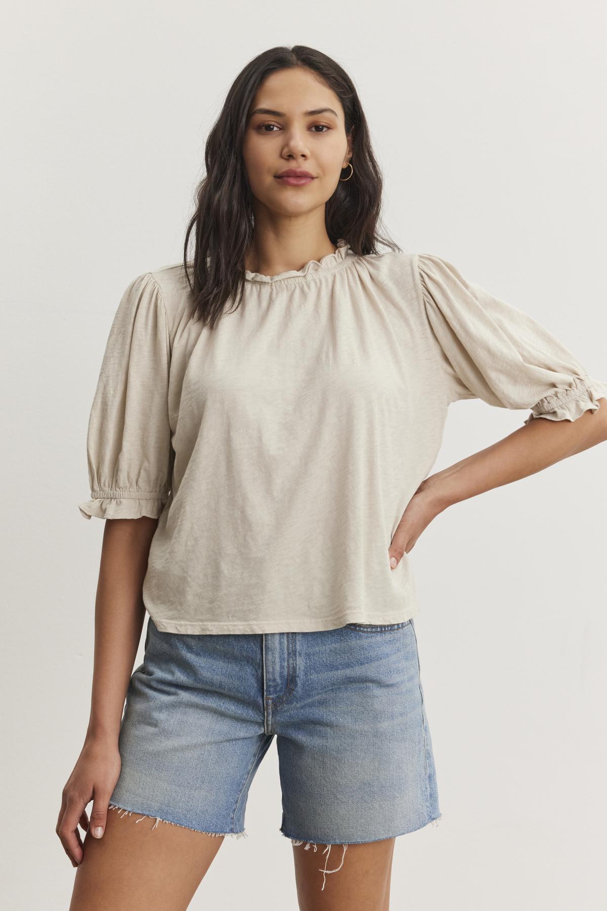   A person with long dark hair wearing a relaxed fit beige Velvet by Graham & Spencer CARLENE TEE with puff sleeves and denim shorts stands with one hand on hip against a white background. 