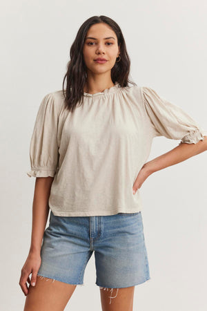 A person with long dark hair wearing a relaxed fit beige Velvet by Graham & Spencer CARLENE TEE with puff sleeves and denim shorts stands with one hand on hip against a white background.