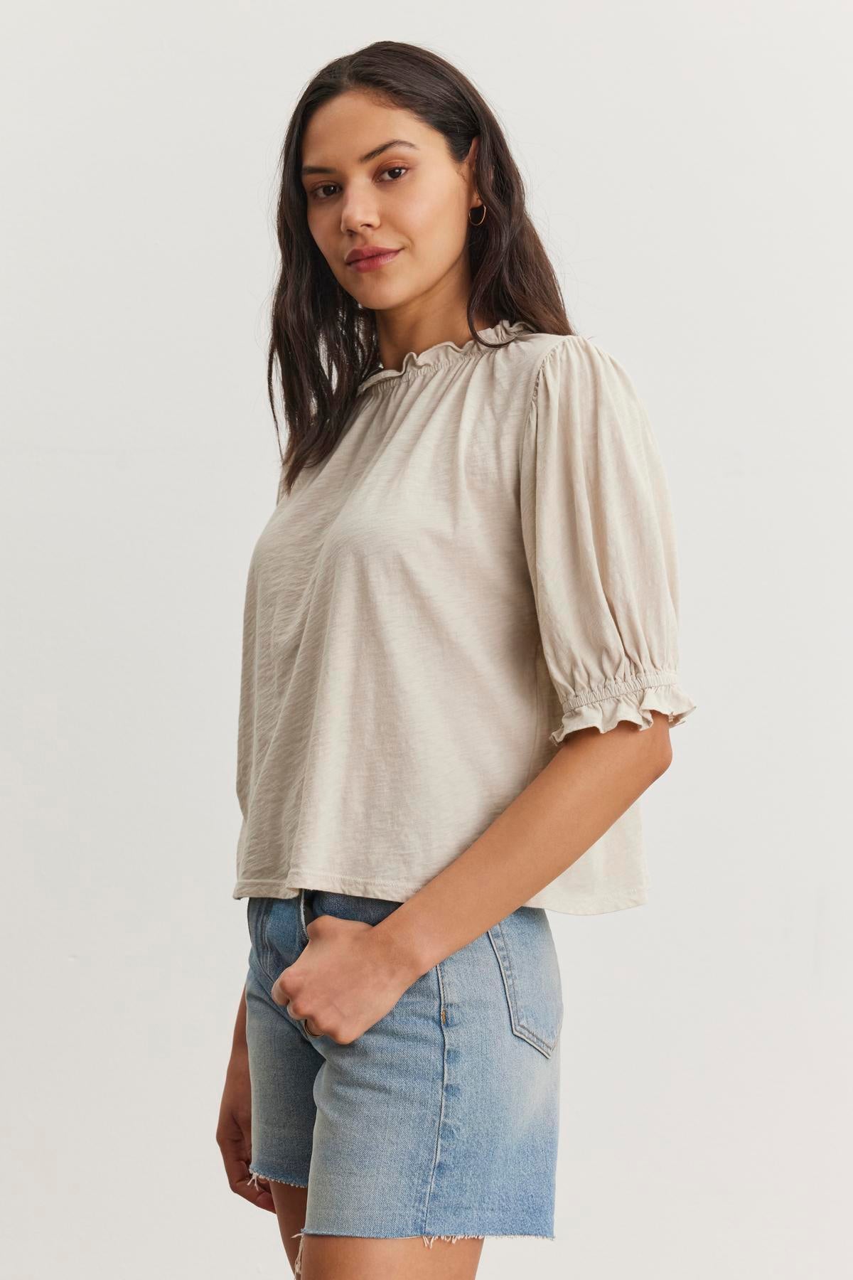   A woman wearing a light-colored CARLENE TEE by Velvet by Graham & Spencer with ruffled sleeves and denim shorts stands facing slightly to the side against a plain white background. The blouse, made of cotton slub fabric, features a relaxed fit that perfectly complements her casual look. 