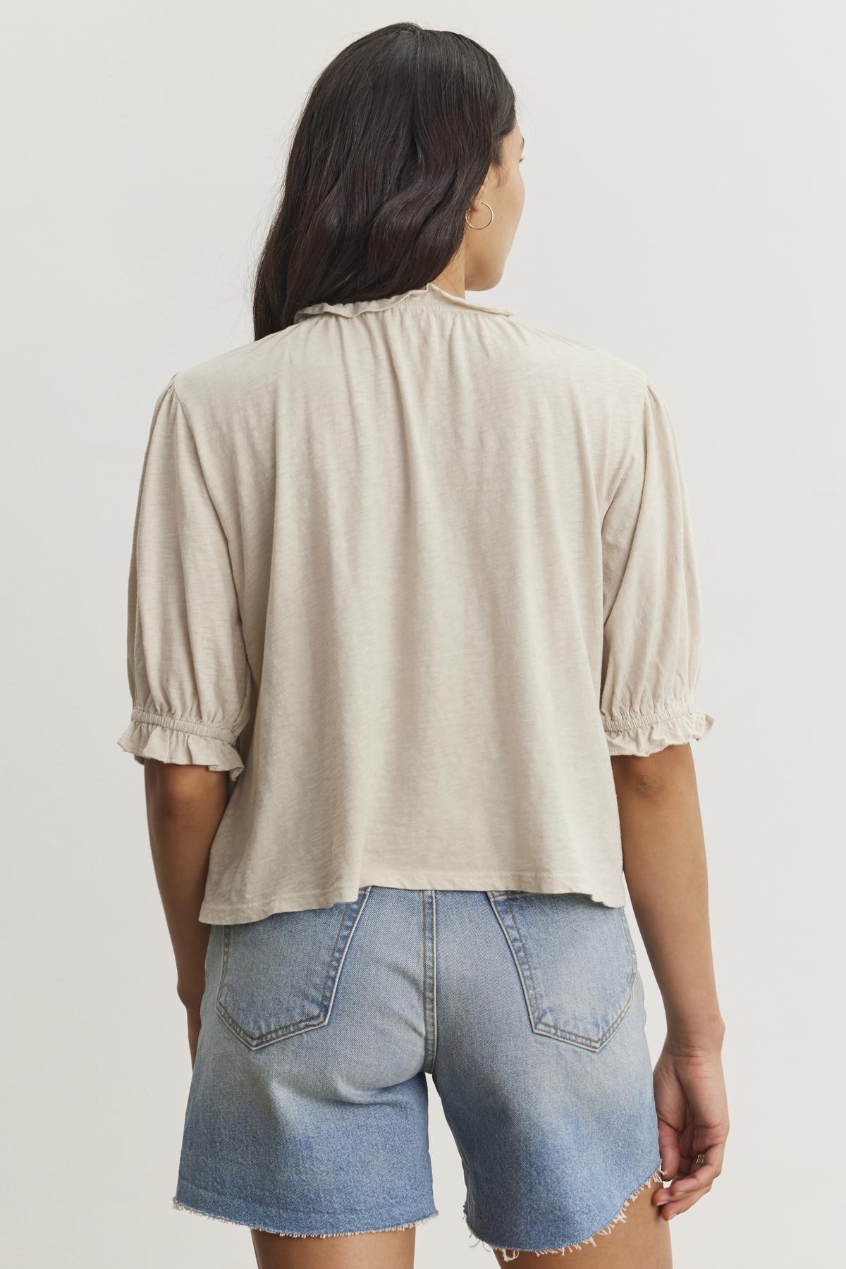   A person with long dark hair is wearing a light beige Velvet by Graham & Spencer CARLENE TEE with a ruffle elastic neckline and blue denim shorts. The relaxed fit of the top and its soft cotton slub fabric add to the overall casual vibe. The person is facing away from the camera, showing the back of their outfit. 