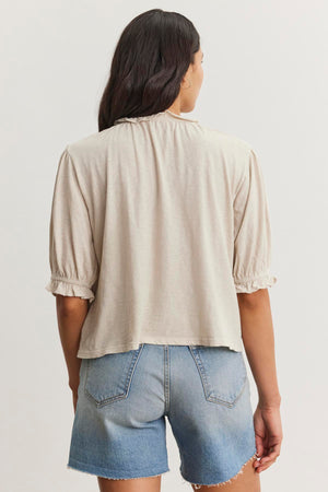 A person with long dark hair is wearing a light beige Velvet by Graham & Spencer CARLENE TEE with a ruffle elastic neckline and blue denim shorts. The relaxed fit of the top and its soft cotton slub fabric add to the overall casual vibe. The person is facing away from the camera, showing the back of their outfit.