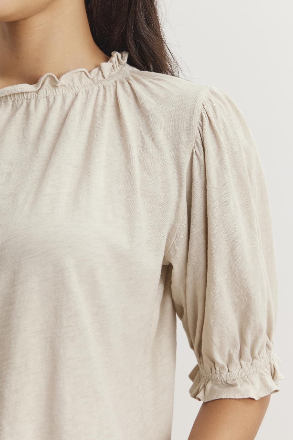   A close-up of a person wearing a light-colored, short-sleeved blouse featuring a ruffle elastic neckline and gathered fabric at the sleeves. The CARLENE TEE by Velvet by Graham & Spencer, made from soft cotton slub fabric, offers a relaxed fit. The background is plain and light. 