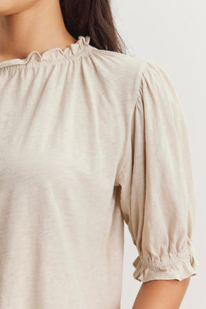 A close-up of a person wearing a light-colored, short-sleeved blouse featuring a ruffle elastic neckline and gathered fabric at the sleeves. The CARLENE TEE by Velvet by Graham & Spencer, made from soft cotton slub fabric, offers a relaxed fit. The background is plain and light.