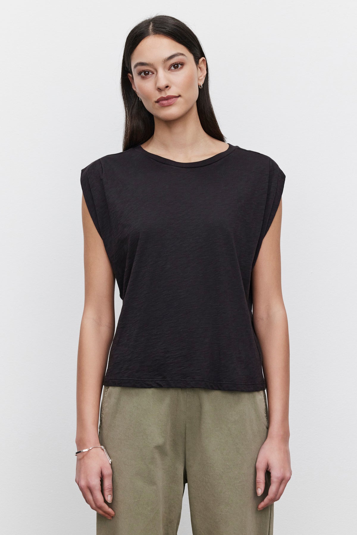   Person with long dark hair wearing a black Velvet by Graham & Spencer Cecile Tank Top with pleated shoulder detail and olive green pants, standing against a plain white background. 