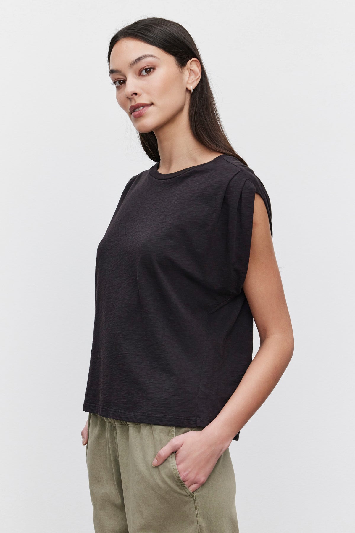 A person with long dark hair wearing a black Velvet by Graham & Spencer CECILE TANK TOP with pleated shoulder detail and light green pants stands against a plain background, looking off to the side.-37629534994625